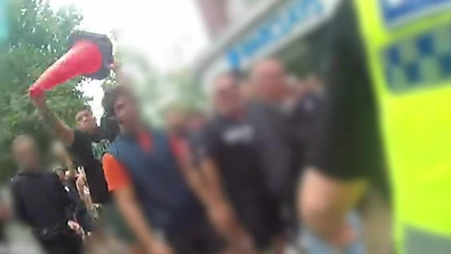 Lewis Carver, wearing a black and green T-shirt, holds a bright red traffic cone above his head, shortly before throwing it at a police officer.