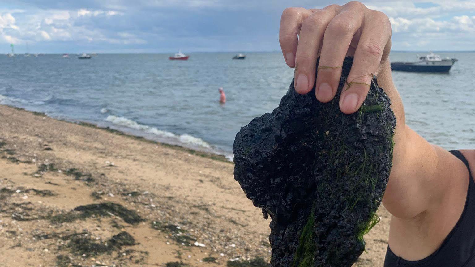 A close up shot of someone's hand holding up a chunk of bitumen, which is mixed with seaweed. They are on a sandy beach next to the sea. You can see one man standing in the sea and a few boats on the water.
