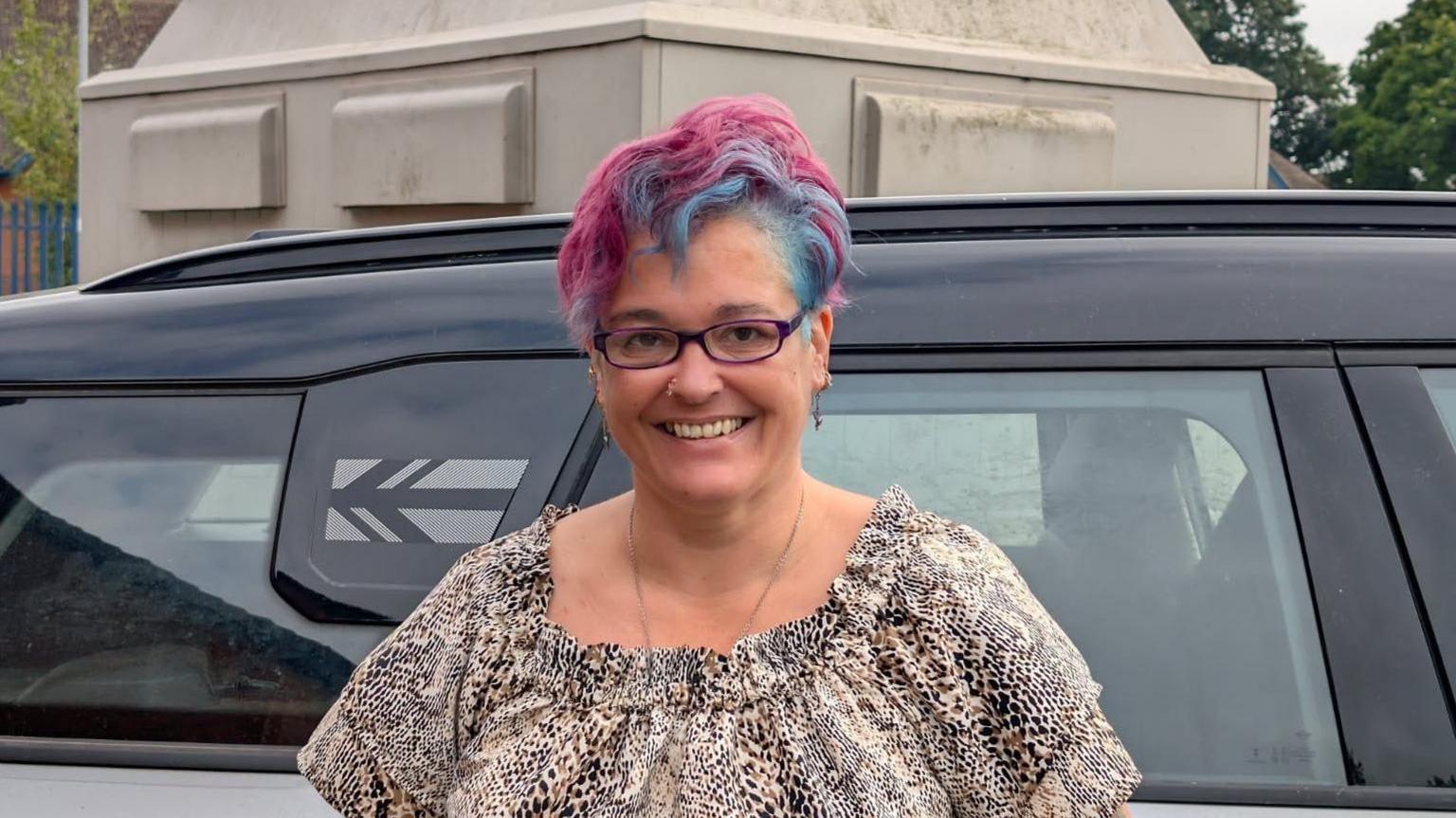 Alison Pope stands against a black car, she is wearing glasses and has a leopard print top. Her hair is dyed blue and pink.  