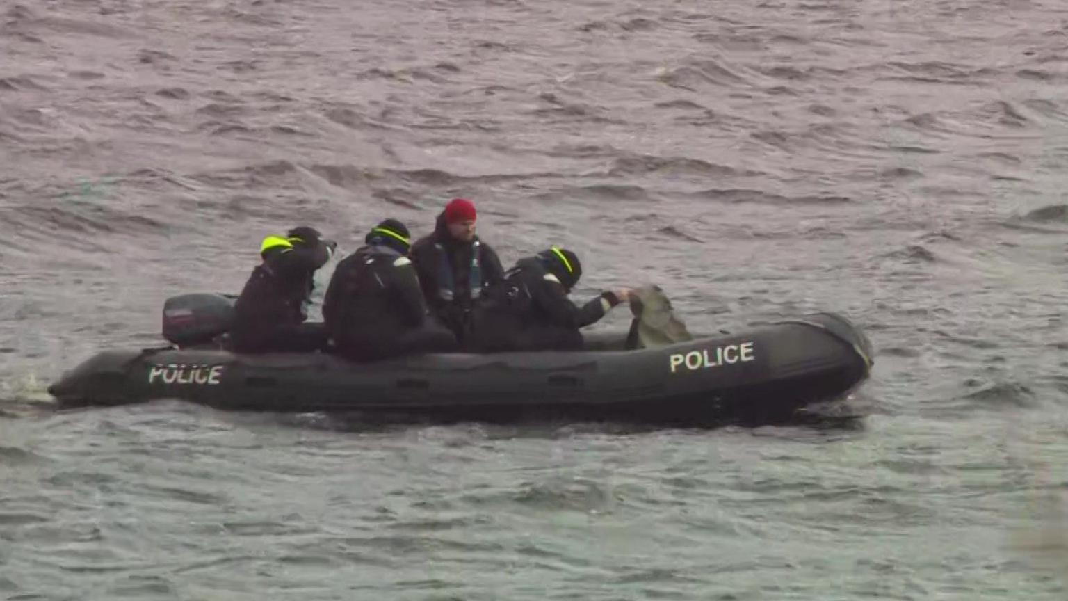 Four men in black wetsuits and hats in a black police dingy on Dovestone reservoir. 