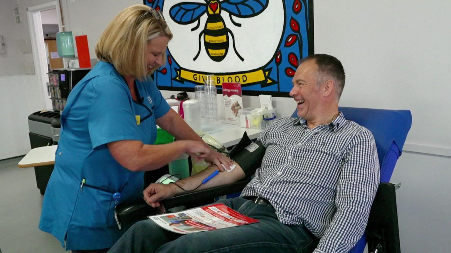 Roger Johnson, who has short hair and is sitting on a blue chair in a medical room, is giving blood. A nurse with blonde bobbed hair and wearing a light blue uniform is collecting blood from Roger's arm. They are both smiling.