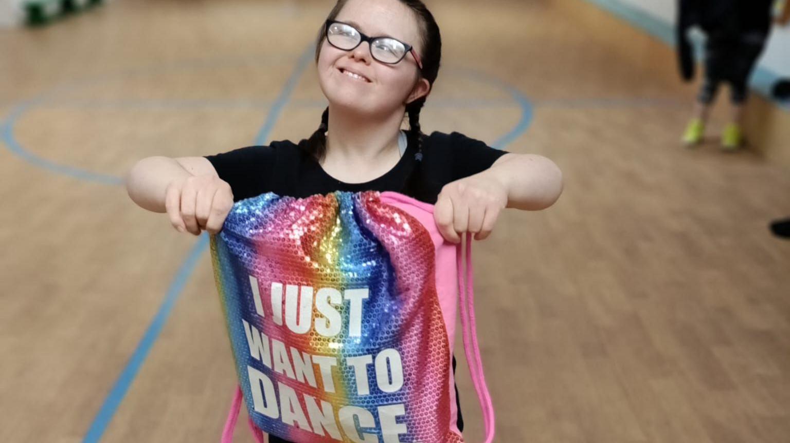 Ms Iles smiles at the camera. She has two hair braids, a black top and is holding up a multi-coloured bag that says 'I just want to dance' written on it.
