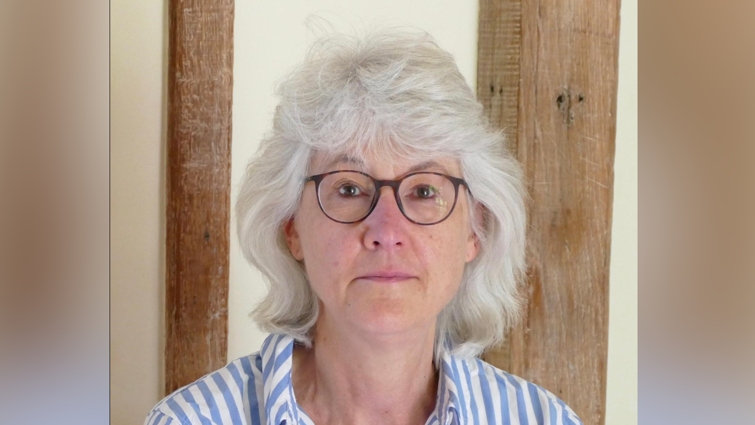 Nicola Rawcliffe smiles at the camera. She has thick grey hair that comes down to the tops of her shoulders. She wears glasses and a white and light blue striped shirt with a collar.