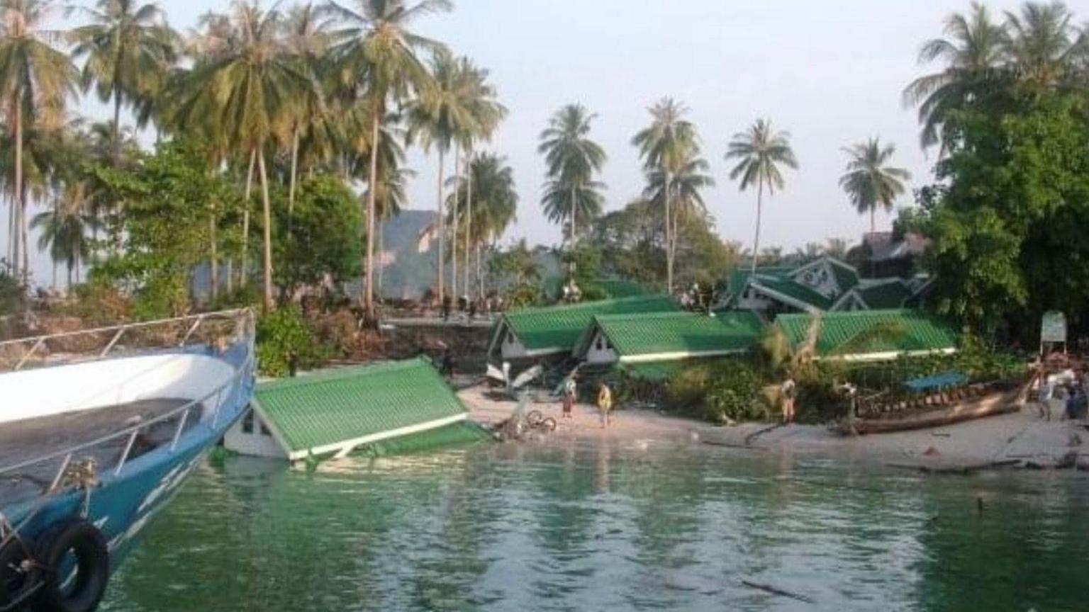 Green and white resort buildings that have collapsed and fallen into the sea. The tide level is still very high in the aftermath of the tsunami. There are people on the beach assessing the damage. In the background there are lots of palm trees still standing.