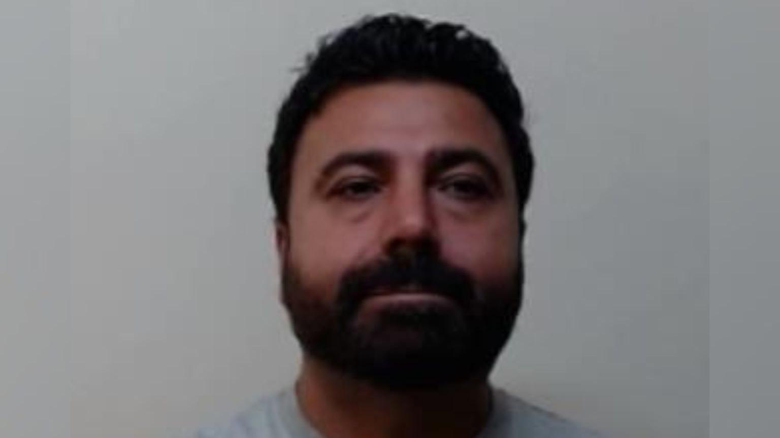 A mugshot of Kenan Baki. He has dark hair and a dark beard and is staring straight at the camera. He is wearing a grey jumper and is standing against a grey background.