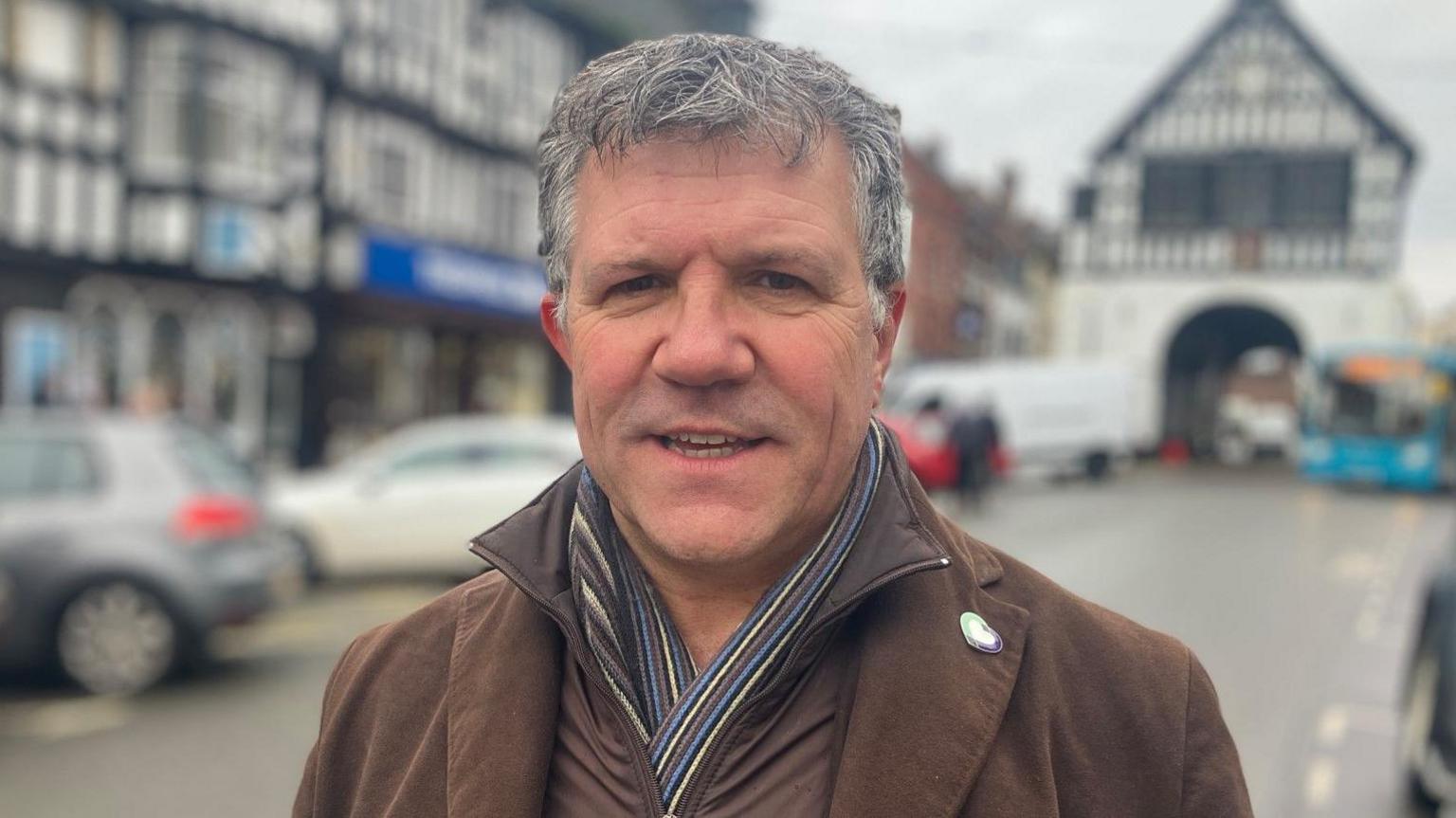 Johnny Themans with grey hair has a brown coat and a scarf. Cars are parked in front of buildings in the background.