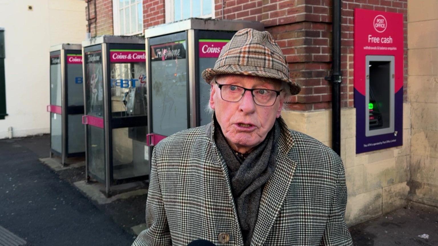 David Medina stands outside the post office. There are three BT phone booths behind him to his left and the Post Office cash machine behind him to his right. He wears a traditional beige, red and cream tartan print trilby hat with a matching smart tartan woolen coat and a brown woolen scarf around his neck. Mr Medina wears thin black framed glasses and has white hair.