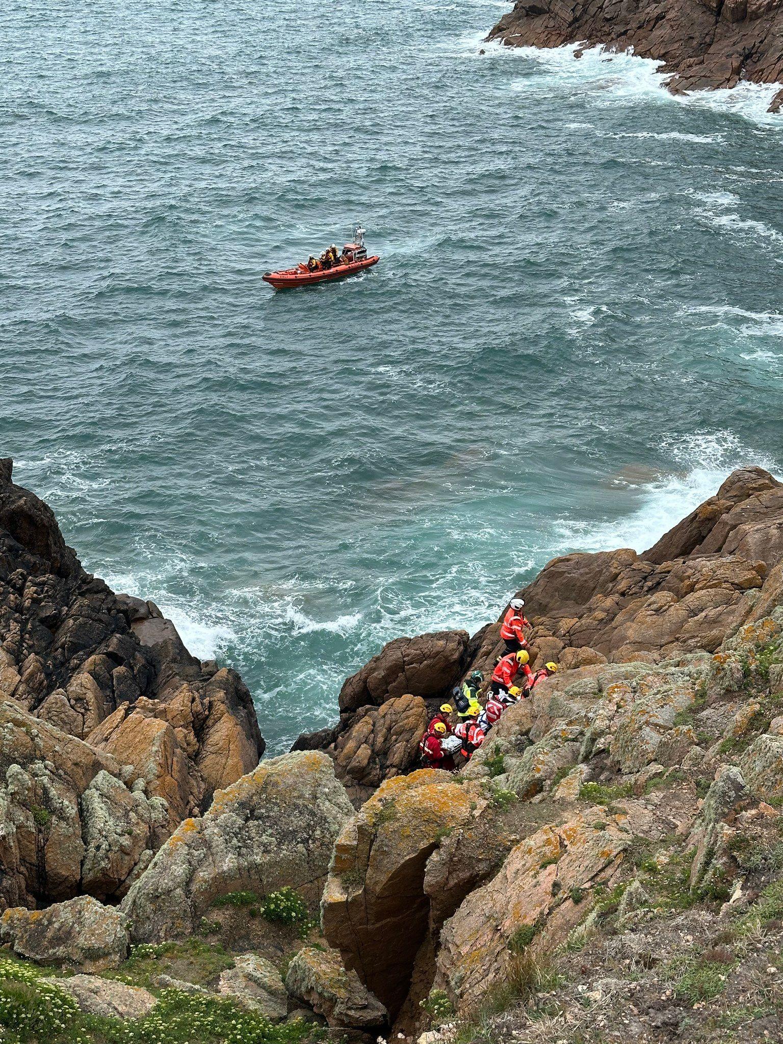 A wider shot of the rescue from the top of the cliff. Nine emergency service workers wearing high-viz uniform and helmets are grouped together in a gap between some rocks. The cliffside is steep and rocky. There is a lifeboat in the bay and the sea looks choppy.