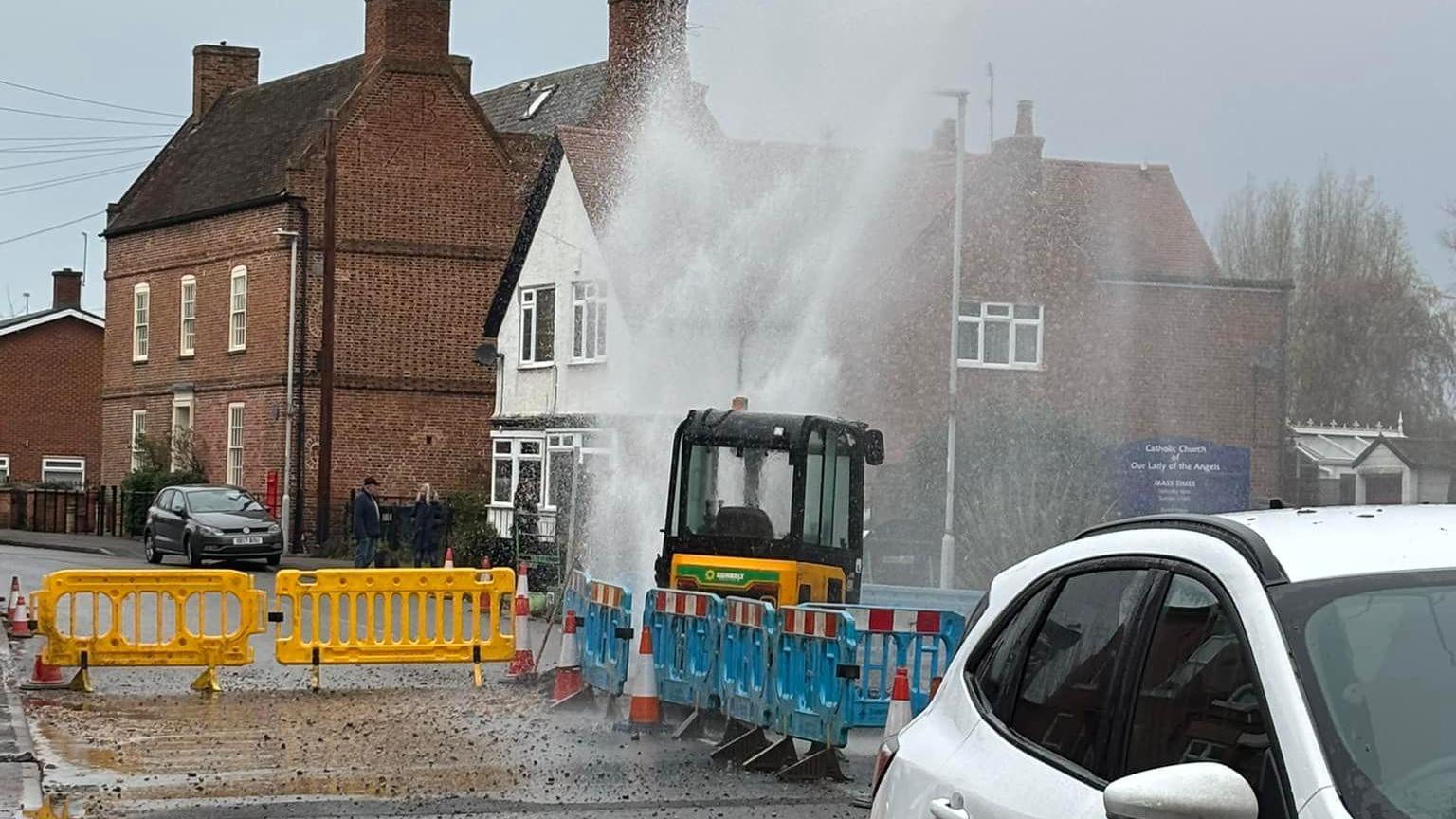 A photograph of water bursting out of the ground in the middle of a road. Around it are temporary barriers and a piece of machinery working on the repair