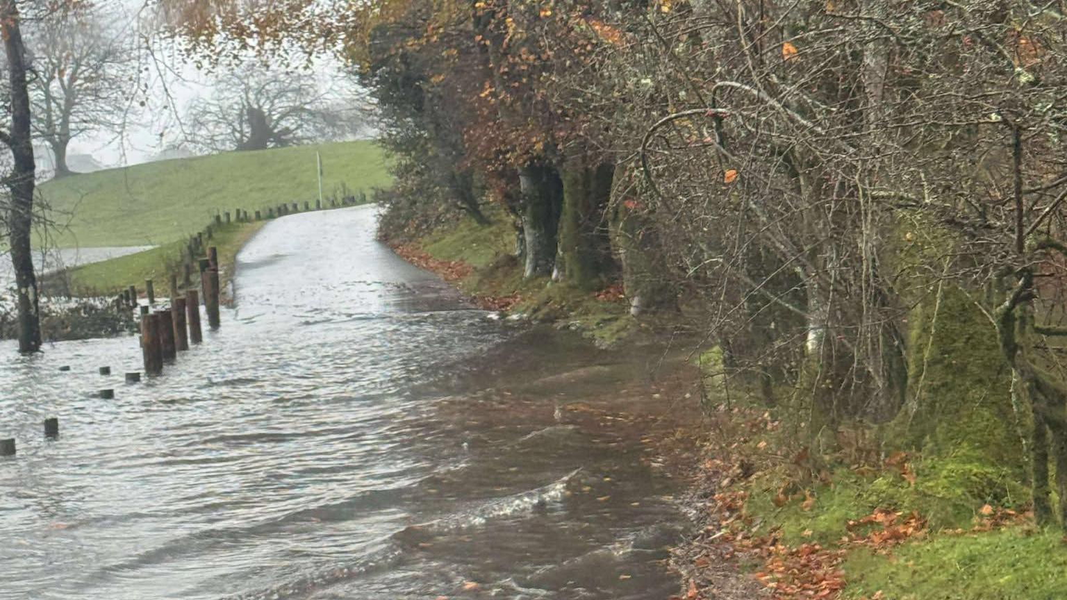 A small road/track on the western shore of Windermere. Flooding has covered the track, which has risen above the fencing. It is a gloomy, wet day with hundreds of brown leaves scattered on the ground.