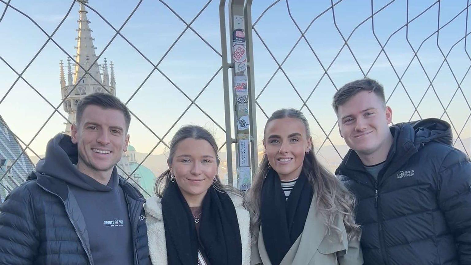 Dion, his partner and their two friends smile looking at the camera. They are in a building which is seemingly tall considering the background. You can see the top of a building that could be a church or a cathedral.