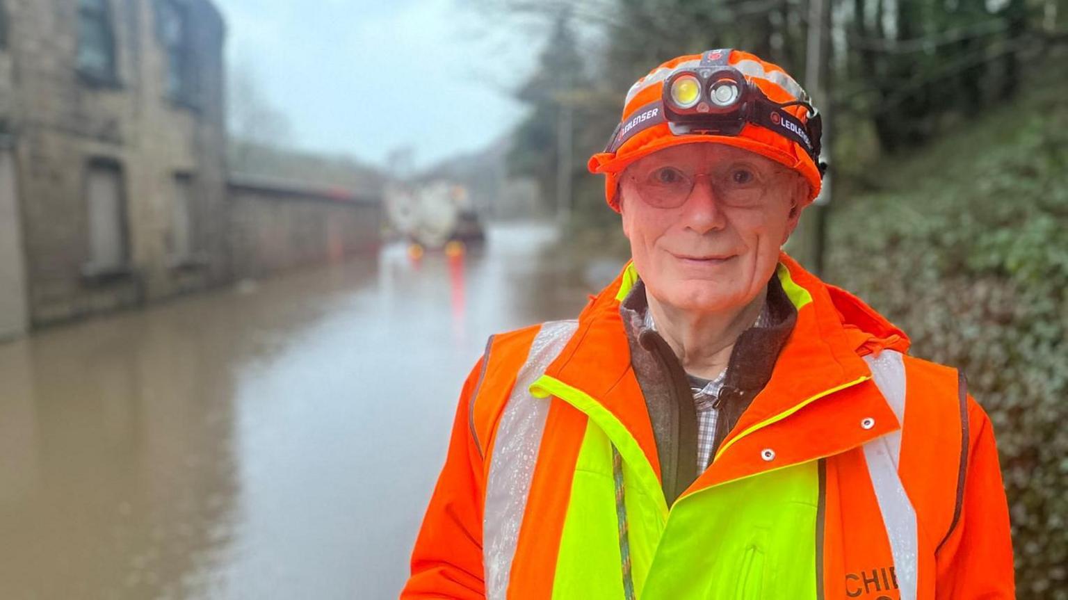 A man wearing luminous orange and yellow overalls and a hat stands against the backdrop of floodwater along a main road.