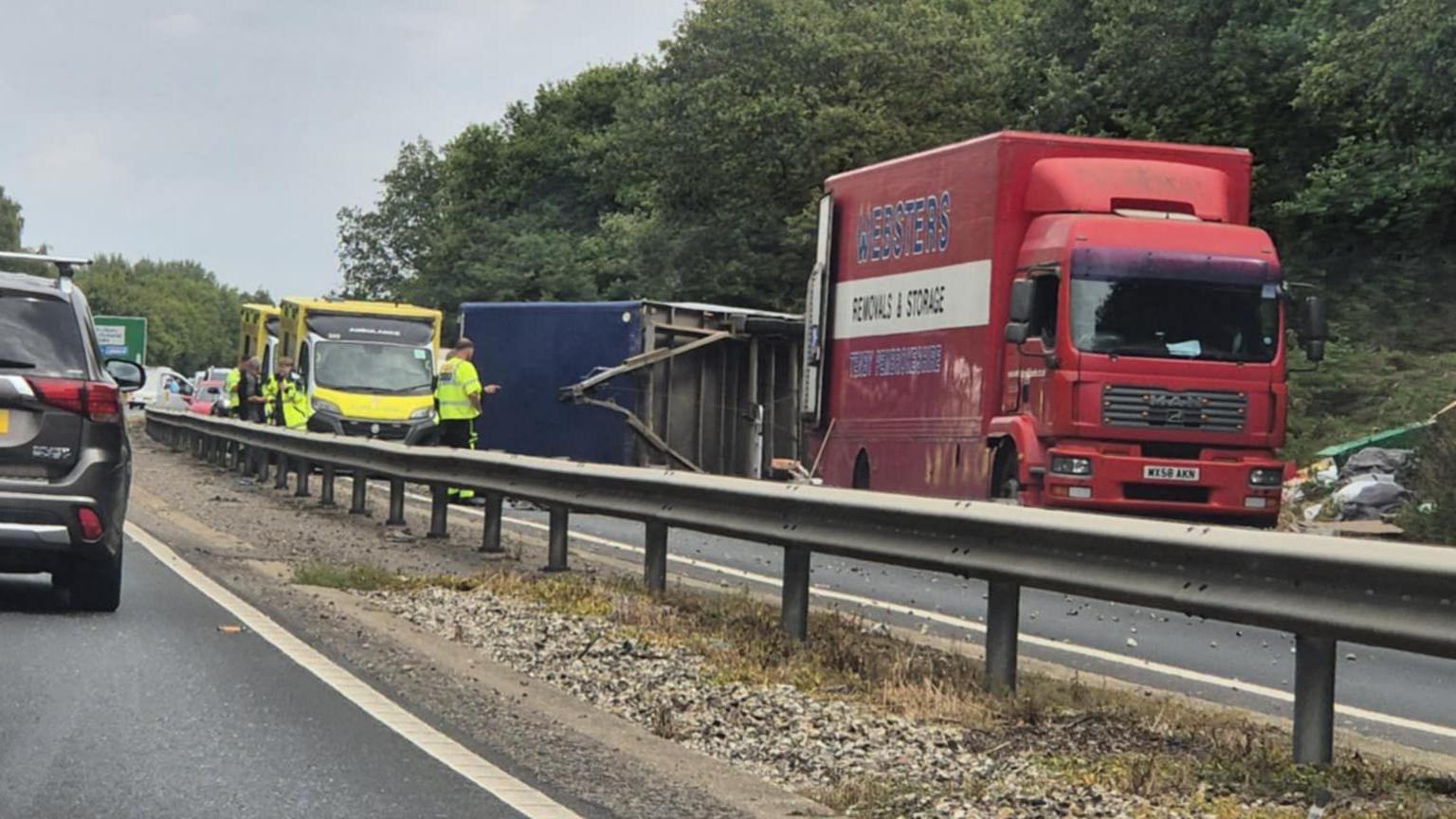 A lorry overturned on the A12. There are police officers standing around the car and there is an ambulance parked.