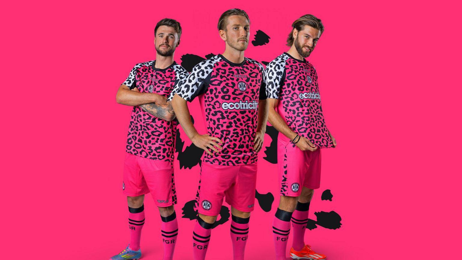 three footballers pose in the away version of the kit which is coloured bright pink.