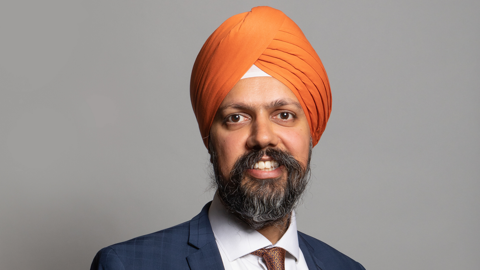 Tan Dhesi's Parliamentary headshot. He is wearing an orange turban and a dark blue suit with a white suit and patterned red tie 