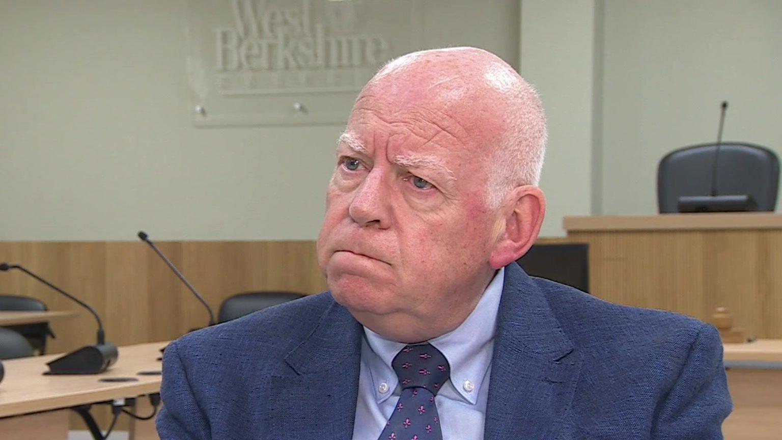 Jeff Brooks frowns during a TV interview in a council chamber. He is balding, with short, white hair and wears a blue jacket, shirt and tie.