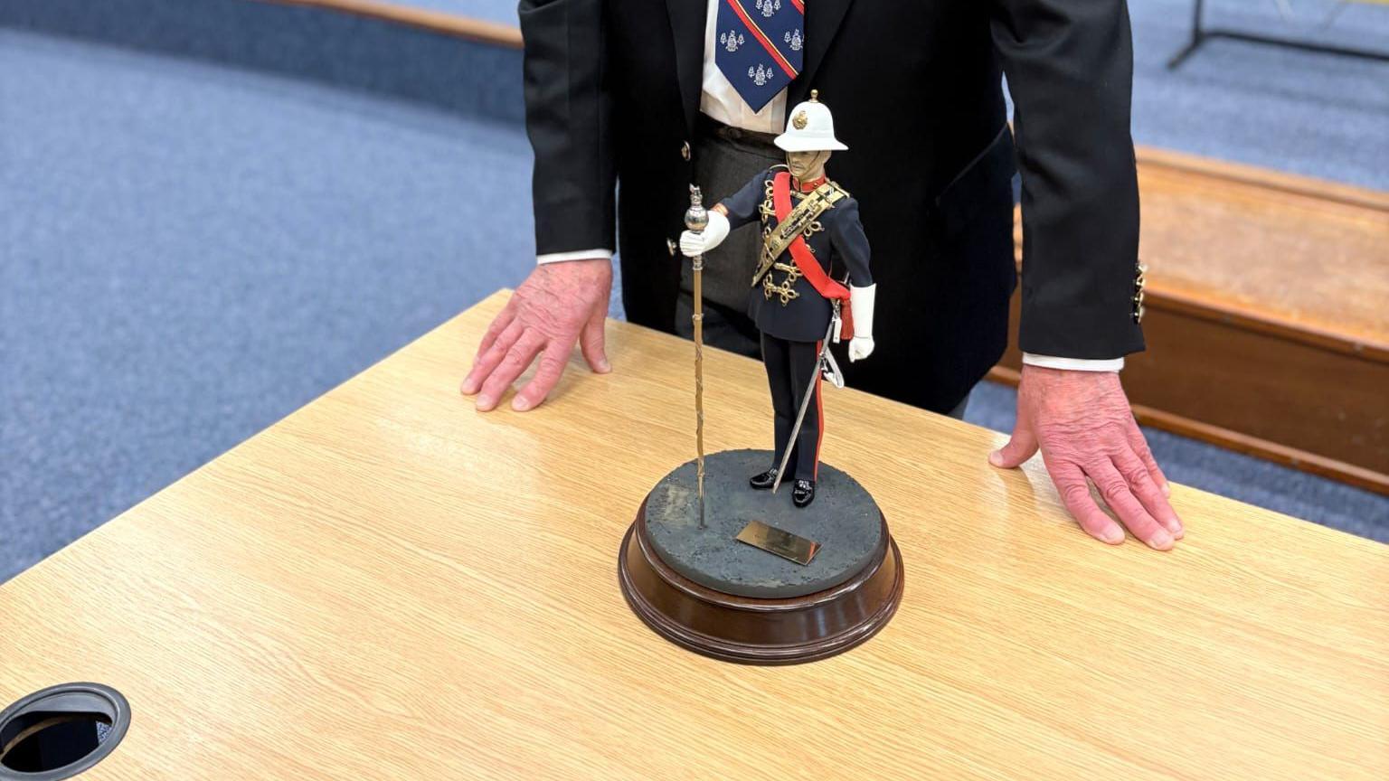 A statue of a drum major which is about eight inches tall. He is wearing a dark blue uniform with a red sash on a wooden plinth.