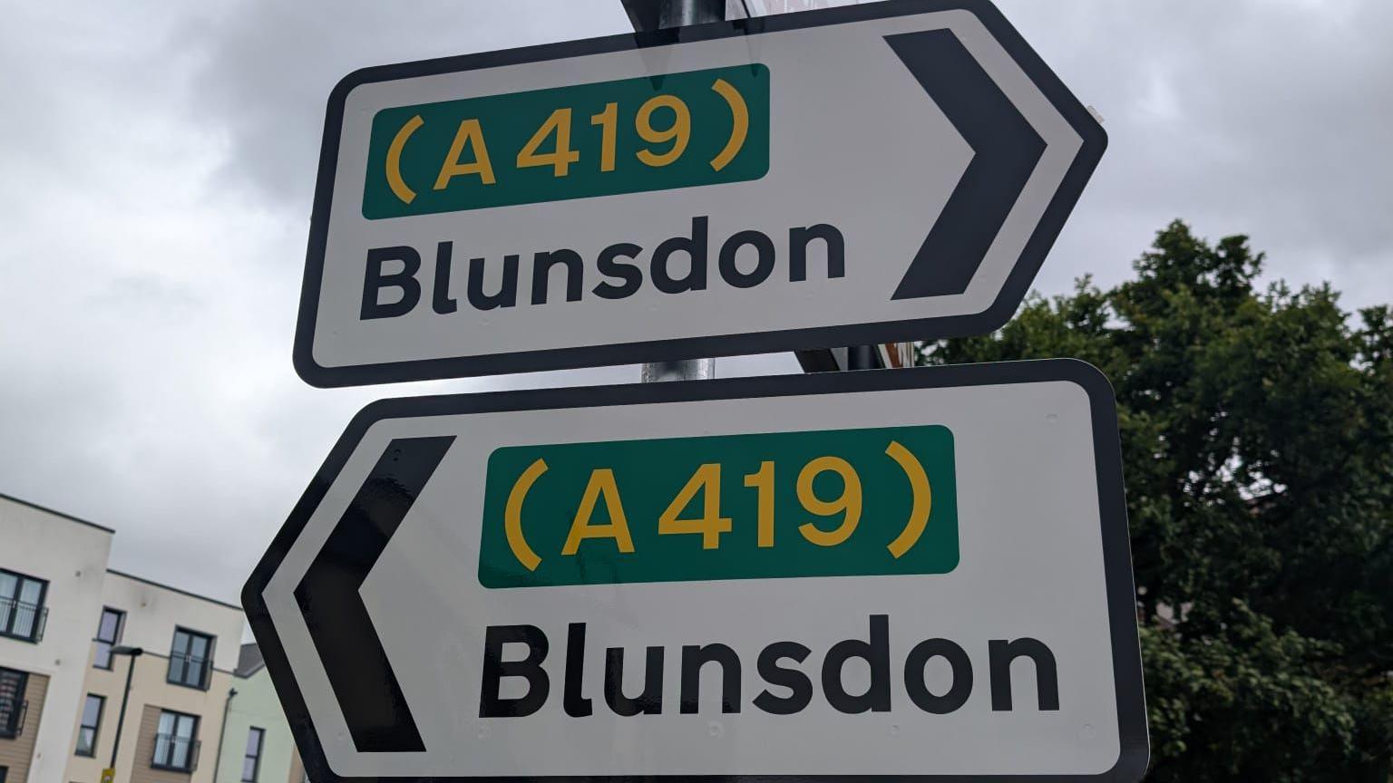 Two smaller official road signs - the upper one points to the right, saying it goes to the A419 and Blunsdon. The one just below points to the left and also says it goes to the A419 and Blunsdon. A grey sky above.