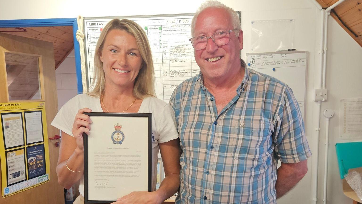 A picture of a man and a woman stood next to each other smiling at the camera. The woman on the left of the image is wearing a necklace, a white shirt and has blonde hair. She is holding a letter in a frame. The man on the right is wearing glasses, has grey hair and a blue pattern shirt.