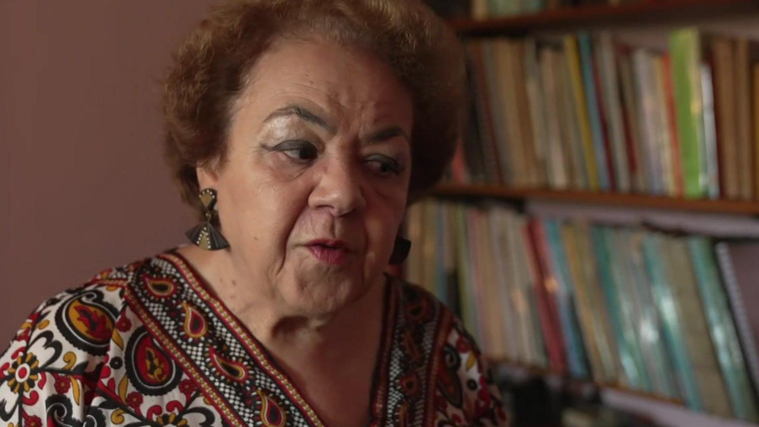 The image shows a person standing in front of a bookshelf filled with various books. They are wearing a colourful patterned top and earrings.