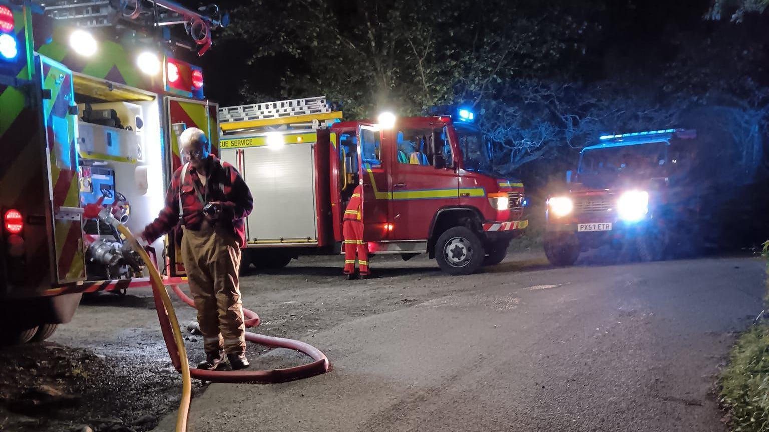 A firefighter holding a hose near a fire engine, with two more fire engines in the background.