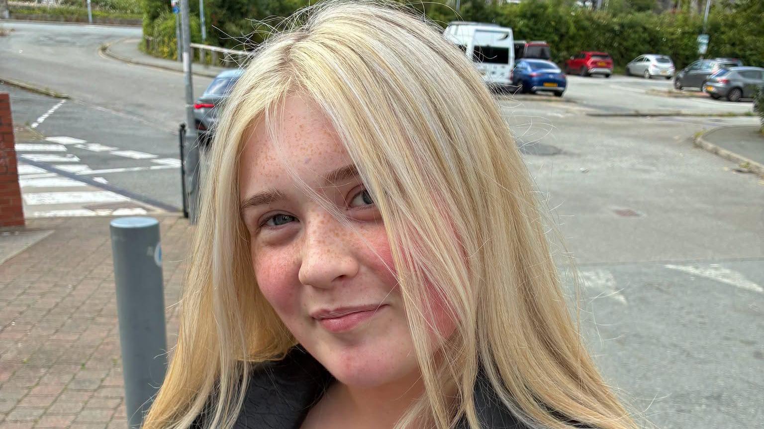 Lucy posing for photo in the street. She is a white women with long blonde hair worn down and freckles on her face.