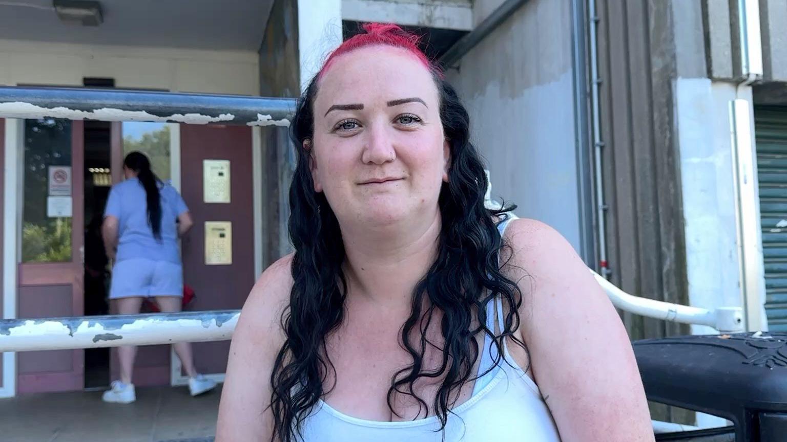 A woman with black and pink hair is looking at the camera. She is stood in front of a block of flats with a door behind her.
