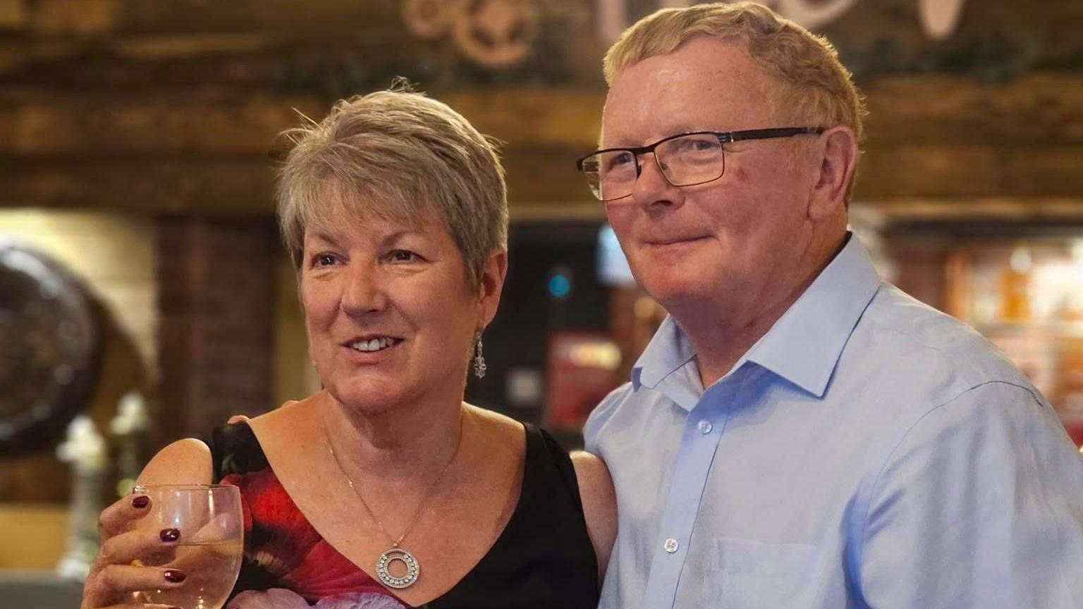 Carol and Keith Fishlock are smartly dressed. Carol has a silver necklace on and a glass in one hand. Keith is wearing a blue shirt and glasses.
