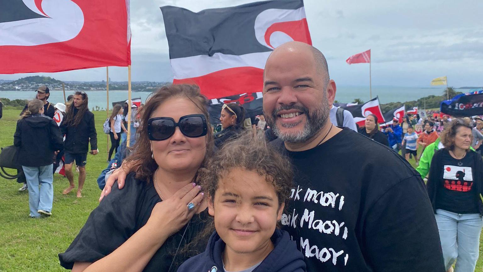 A family of three smiles at the camera, while people wave flags in the background