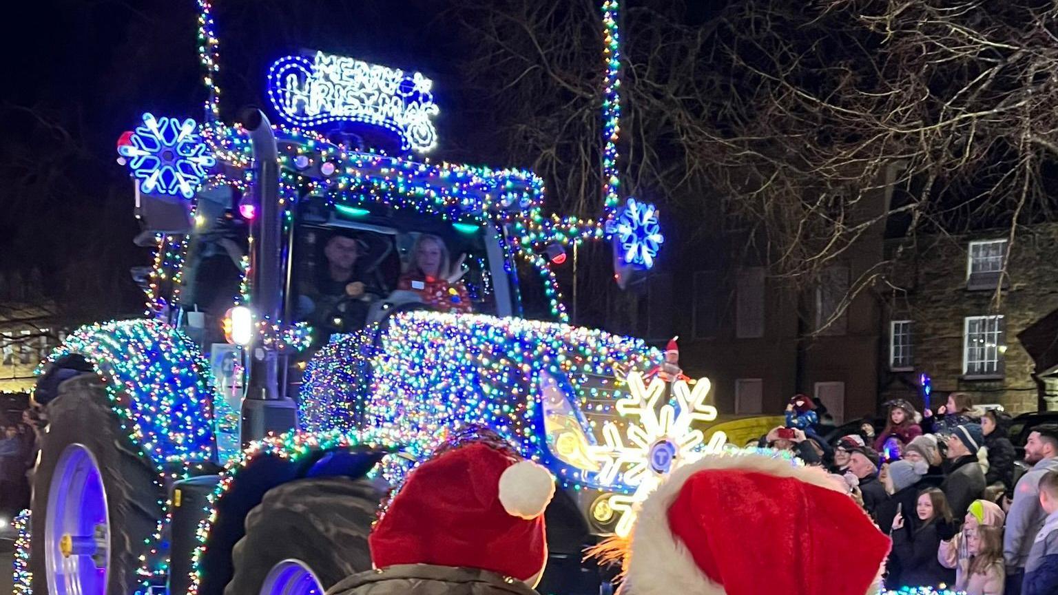 A large tractor lit up with multi-coloured lights. In the foreground are two people wearing Santa hats.