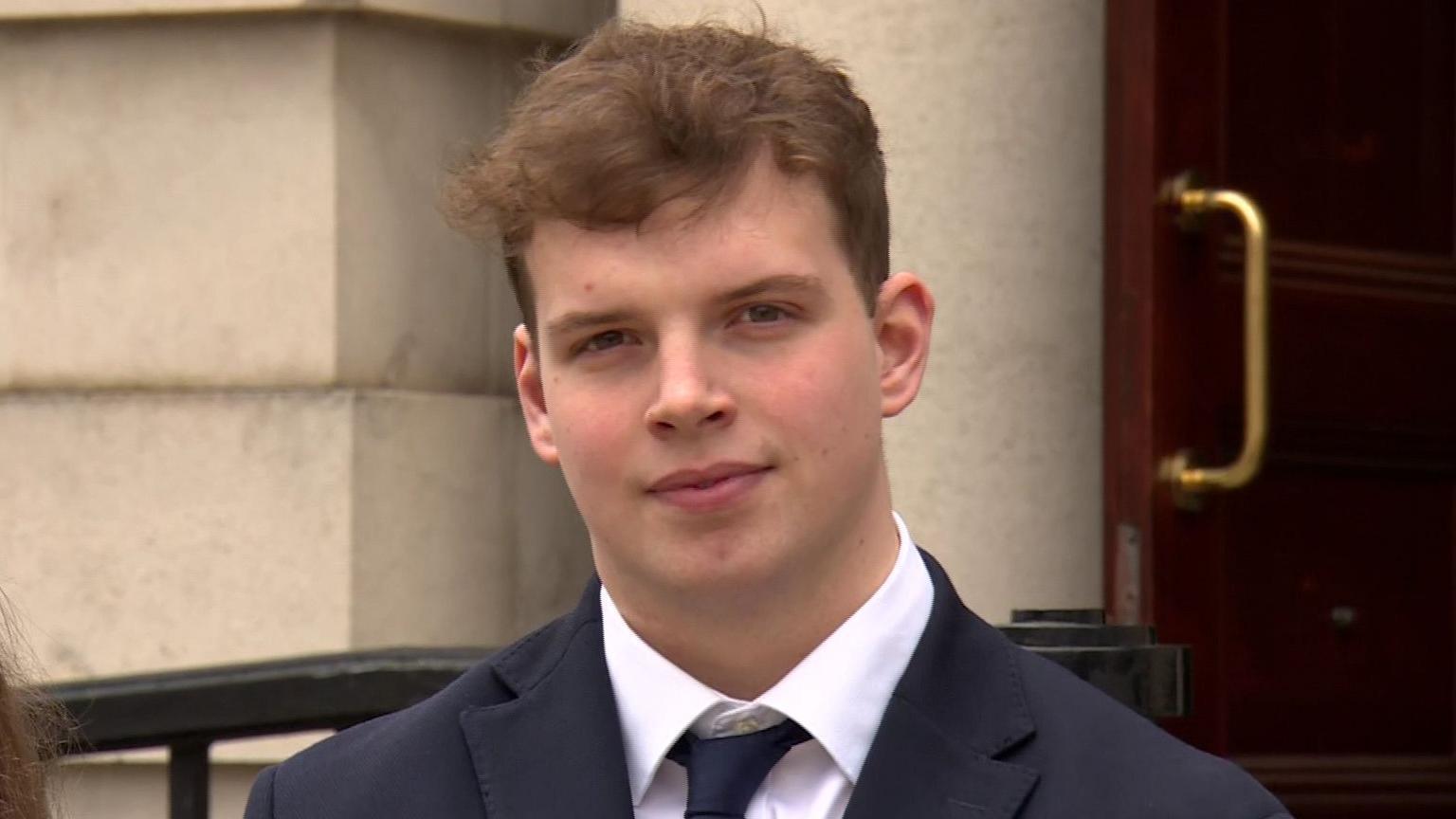 A young man looks into the camera. He's wearing a black suit with a tie and a white shirt. A large building is behind him.