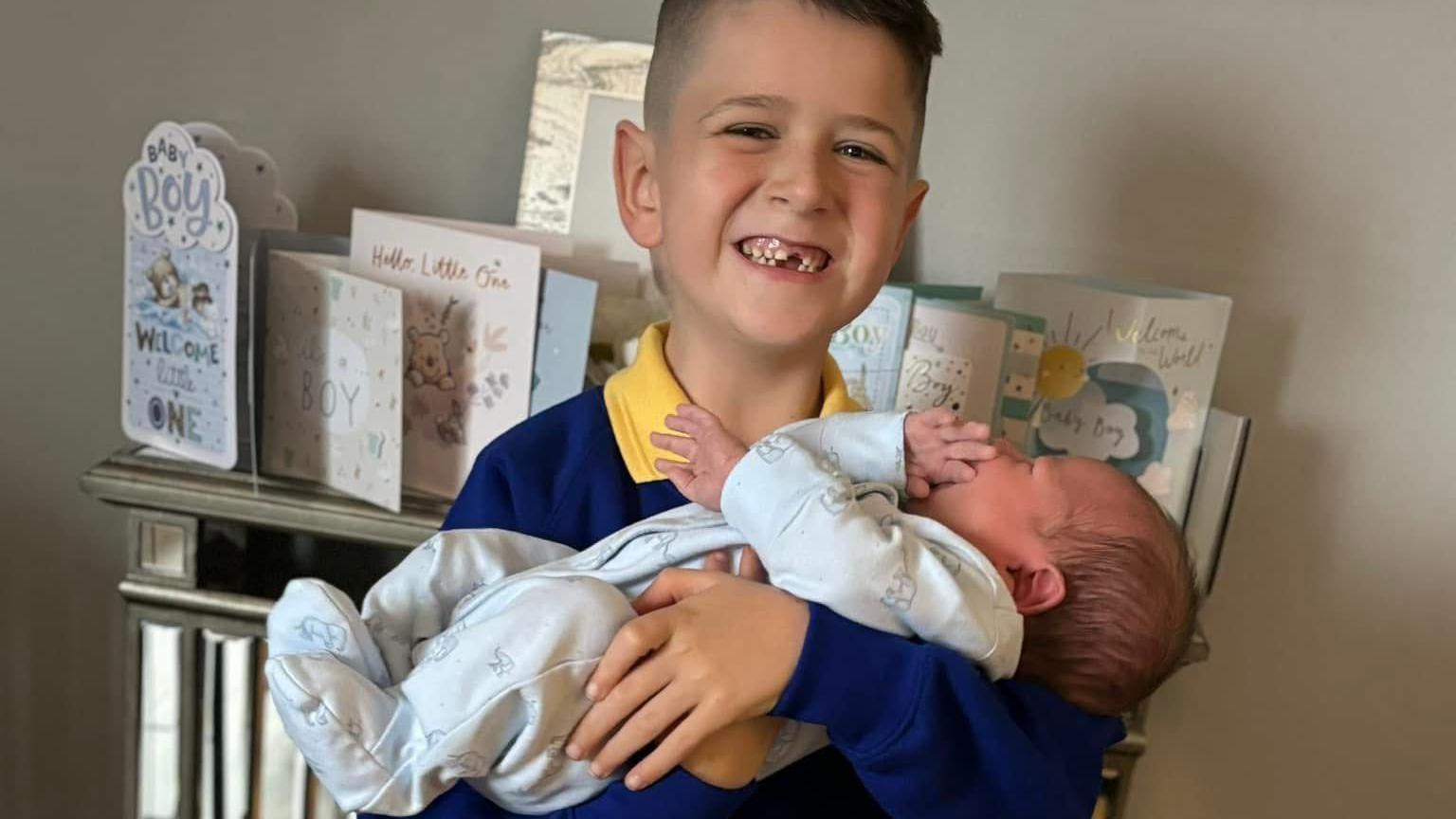 Archie smiles at the camera while holding his baby brother Finley in his arms. Archie has short dark hair and his big grin reveals he is missing his front teeth. He is wearing a school uniform of a yellow polo shirt and blue jumper. Finley has dark hair and is wearing a blue onesie. Behind them is a shelf covered in cards celebrating Finley's birth.