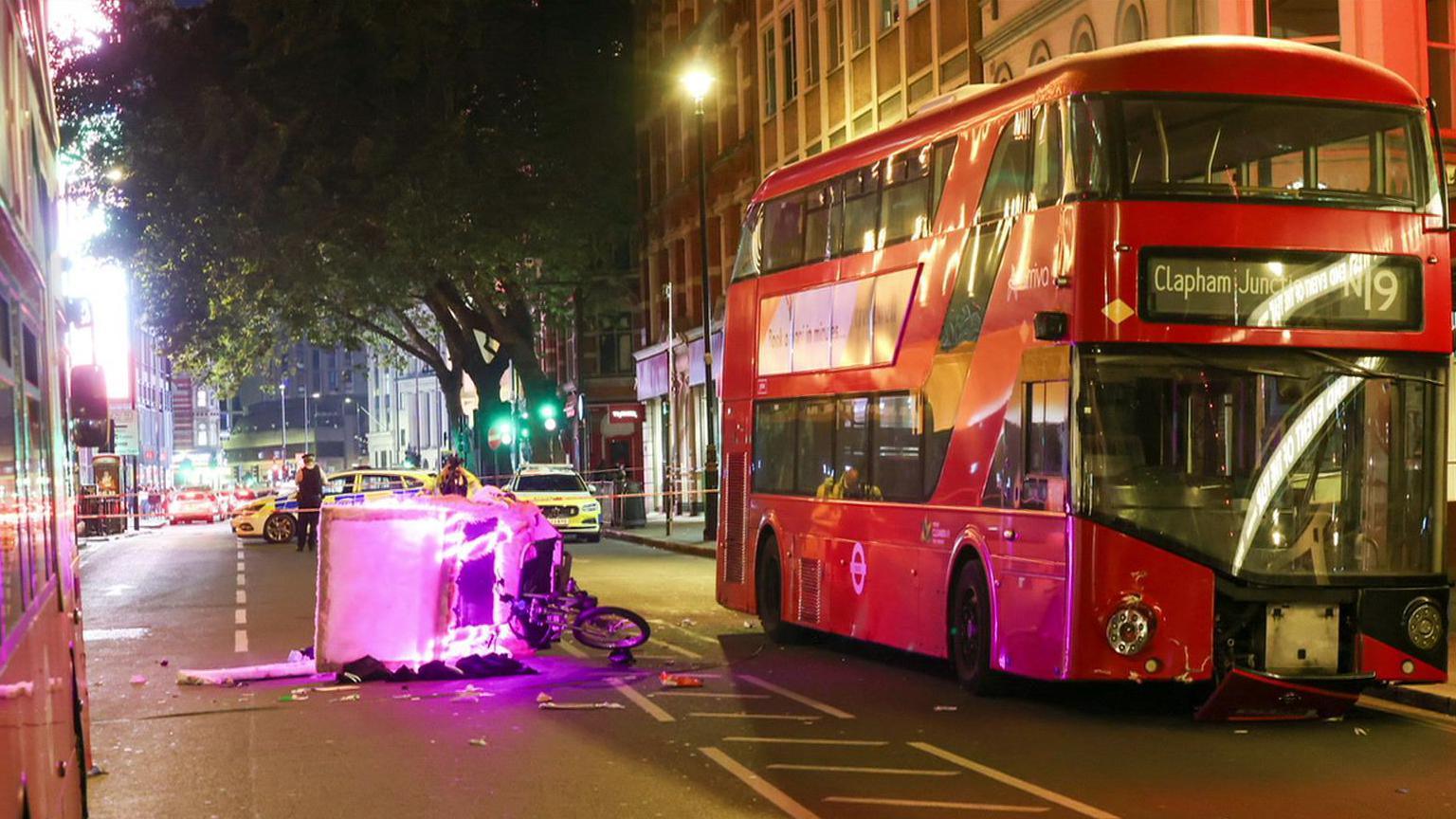 a pink pedicab lies damaged and on its side in the road next to a red London bus as another bus is visible on the other side of the road. Police cars are parked on the road behind and officers are stood in the road. It appears to be night time or the early hours of the morning.