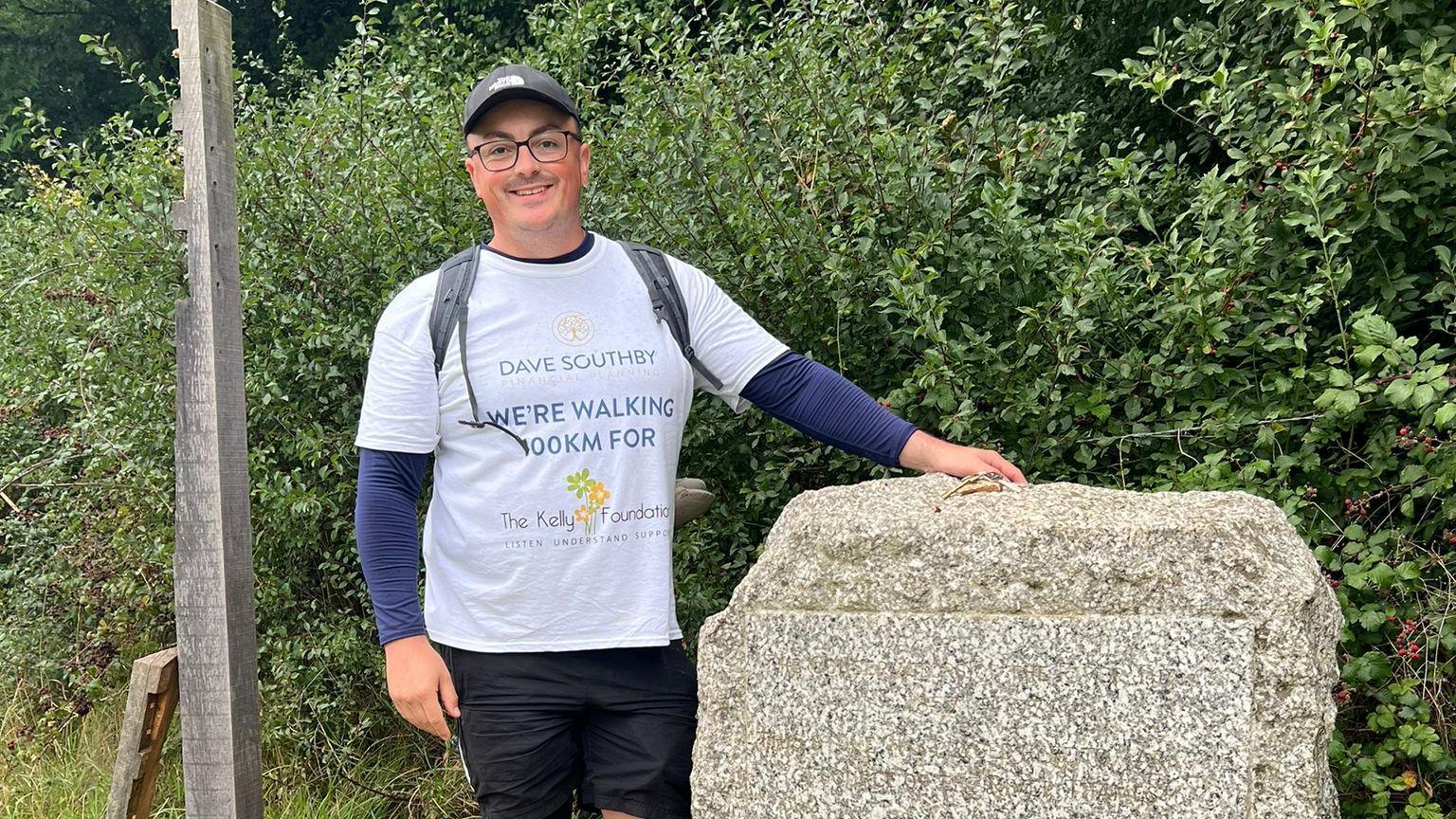 Dave Southby wearing black shorts and a white t-shirt. He is posing and smiling at the camera beside a milestone rock with greenery in the background.