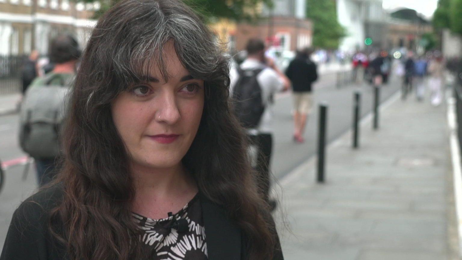 Madeleine Stone is standing in the street, in front of a row of bollards as a line of pedestrians walk past 