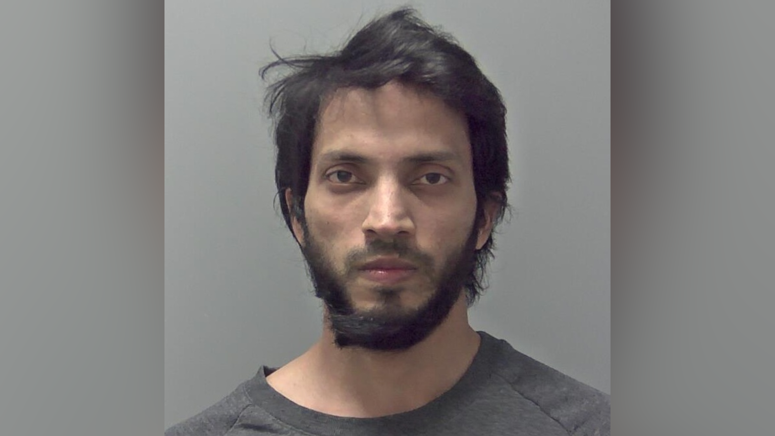 Anwarul Islam's police custody photo. He looks expressionless at the camera. He has medium length black hair and a dark thick beard. He is wearing a grey top. He is standing against a light grey background.