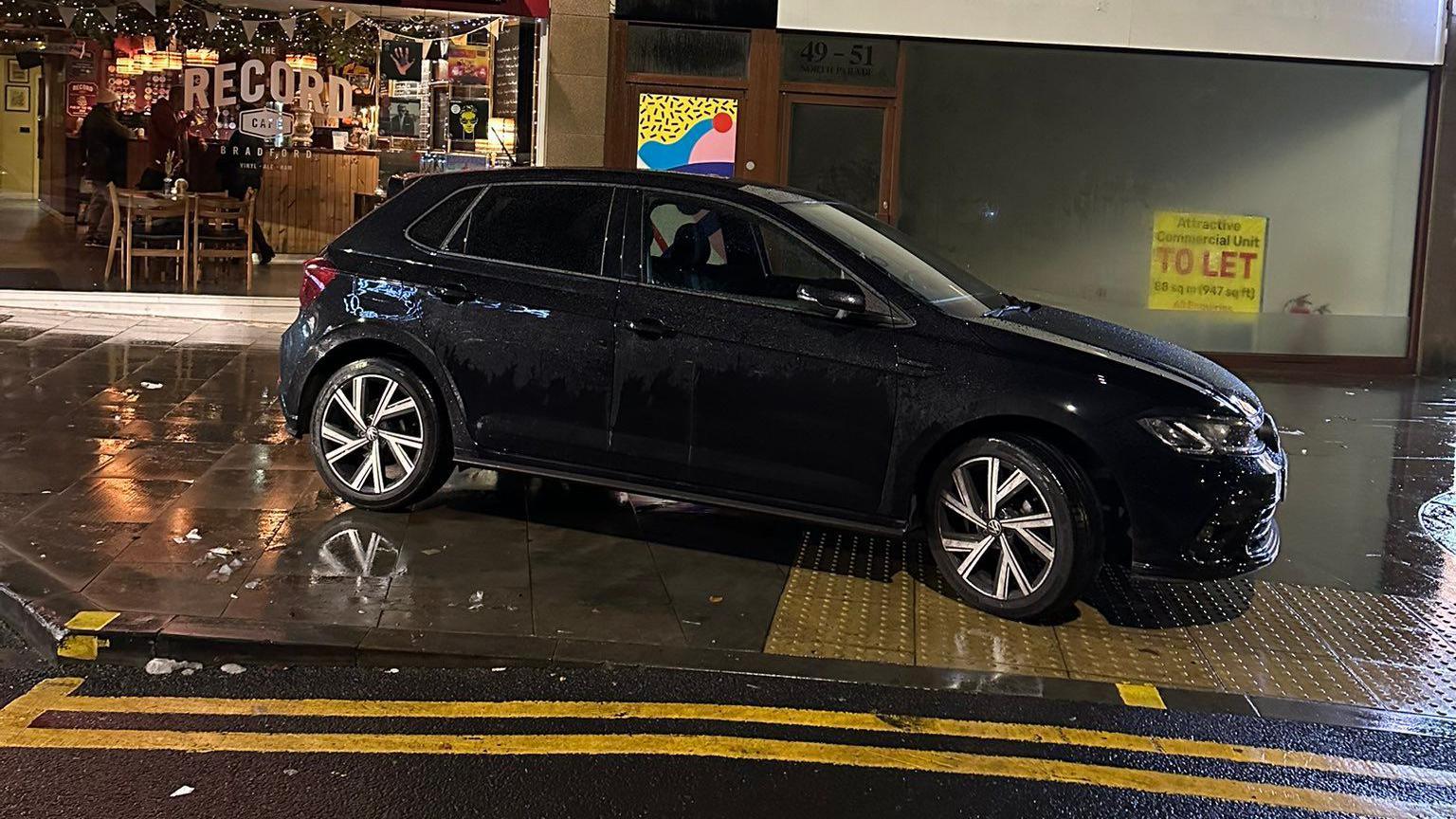 A black hatchback-style car parked on a pavement with double yellow lines in front of it and a bar window in the background.  