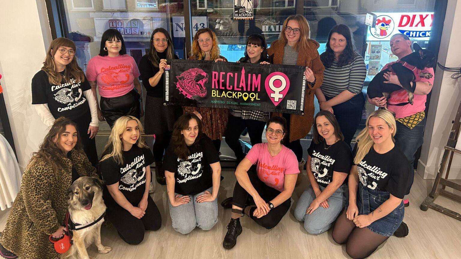 A group of 15 women gather together for a picture, the three in the middle hold a banner that reads 'Reclaim Blackpool' and features a picture of a pink leopard. The women wear pink or black t-shirt that read 'catcalling ain't a compliment' 