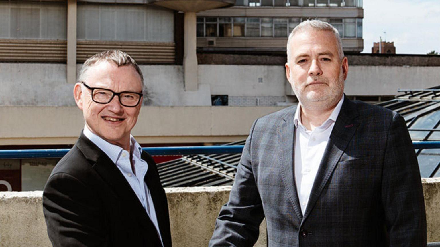 Mike Stonard leader of Norwich City Council is standing next to and shaking hands with Ben Luckett from Aviva. Mr Stonard is wearing a black suit jacket and glasses and is looking directly at the camera and smiling. Mr Luckett is also looking directly at the camera and is wearing a black and grey suit jacket.