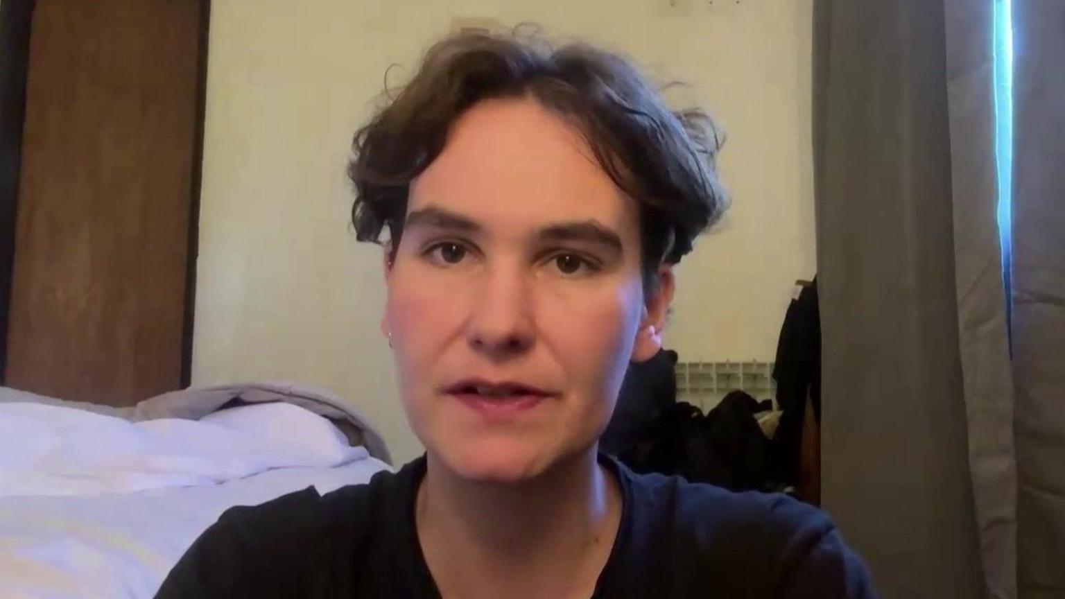 Robyn Lewis is sat down in a hotel room. She is sat next to an unmade bed and a set of grey curtains. She has short brown hair and is wearing a black t-shirt. A door can be seen behind here along with a light yellow wall.