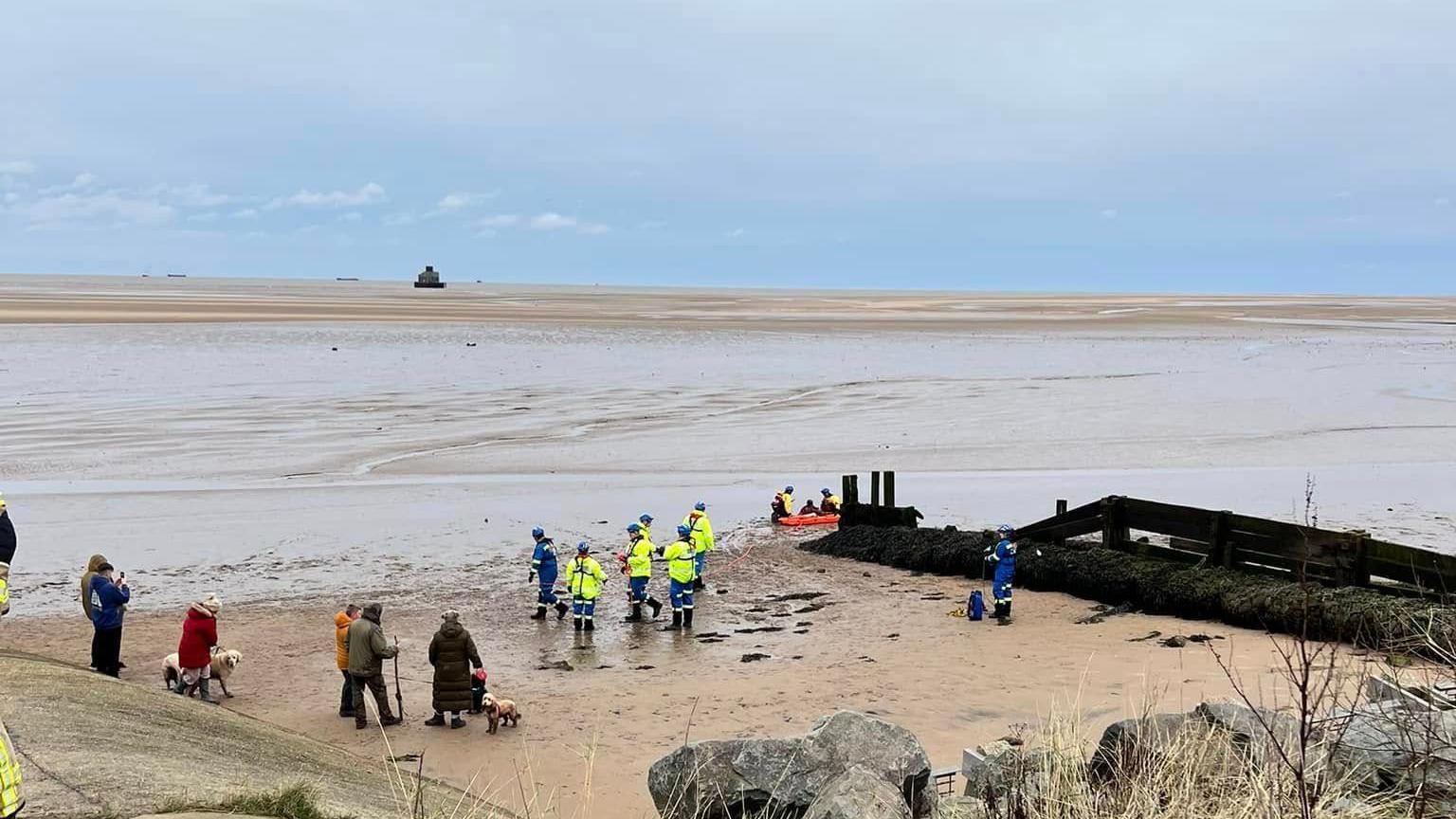 The Humberston Fitties beach with bystanders watching on the sand as a team of emergency workers rescue a man from the mud. A wide expanse of water and sand is visible in the distance as is the Humber estuary which has a number of ships sailing on it.