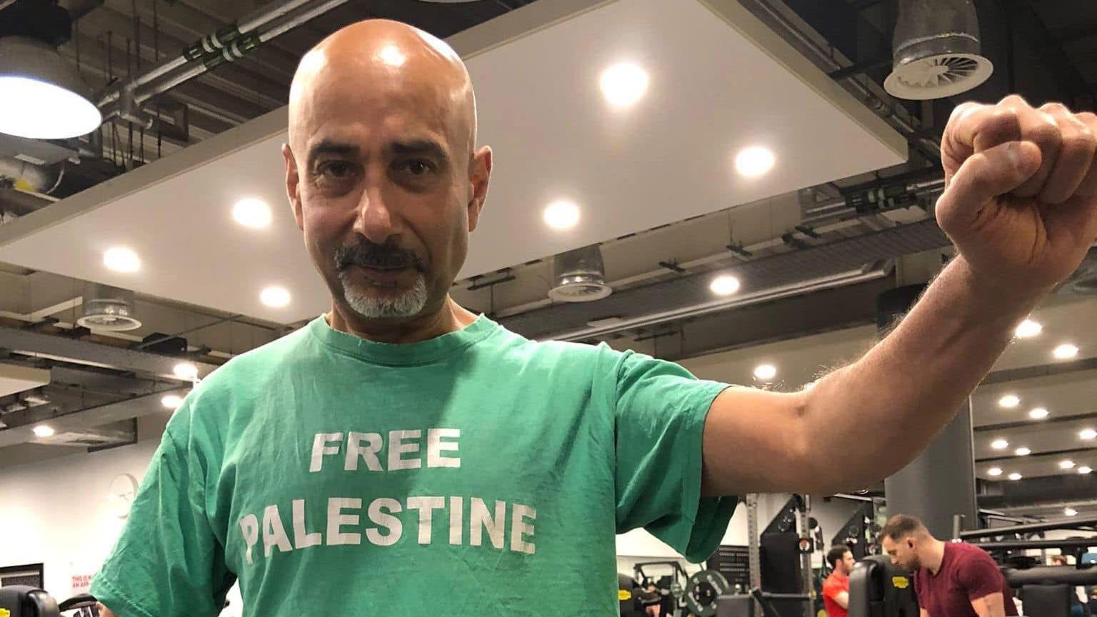 Jasvinder Khosa looks at the camera and holding his clenched fist aloft. He is wearing a green T-shirt with a Free Palestine slogan written in white letter. Behind him several people can been seen working out in the gym.