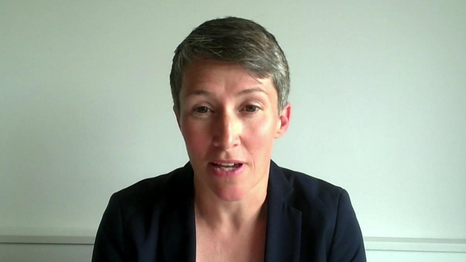 A woman with short hair and a dark top sits in a plane room. She is on a video call and is looking directly at the screen.