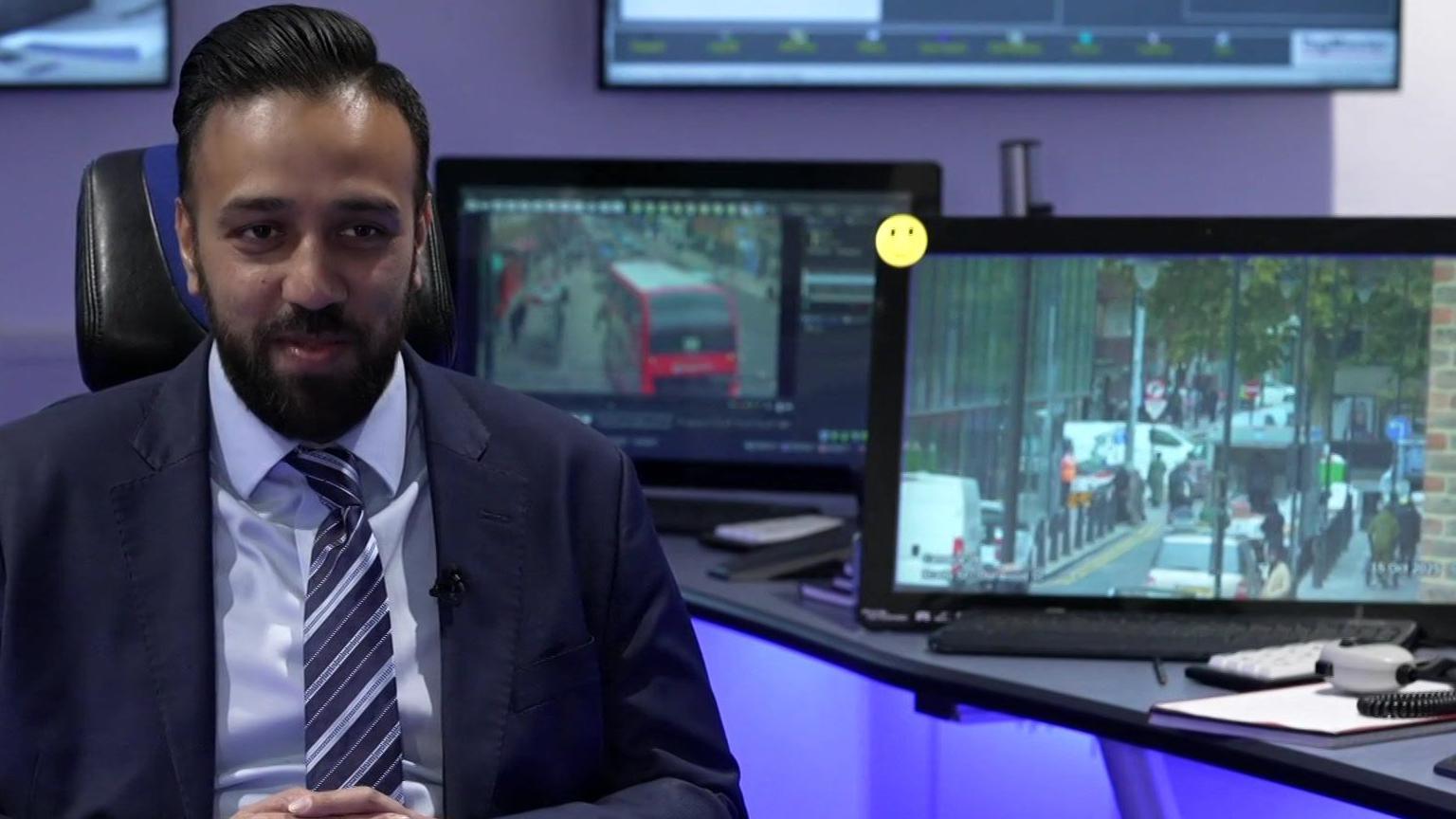 Abu Chowdhury, dressed in a suit and tie, sits in a chair in a room with two computer screens to his left. On the screens are images of local streets from the CCTV cameras