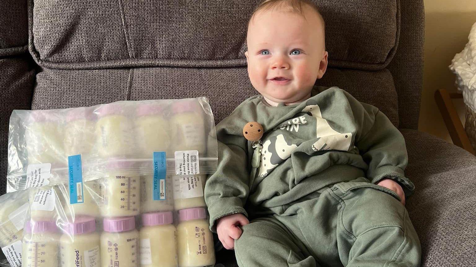 A blond smiling baby boy in a green dinosaur outfit on a grey sofa with a clear bag of thirty or so milk bottles beside him.  