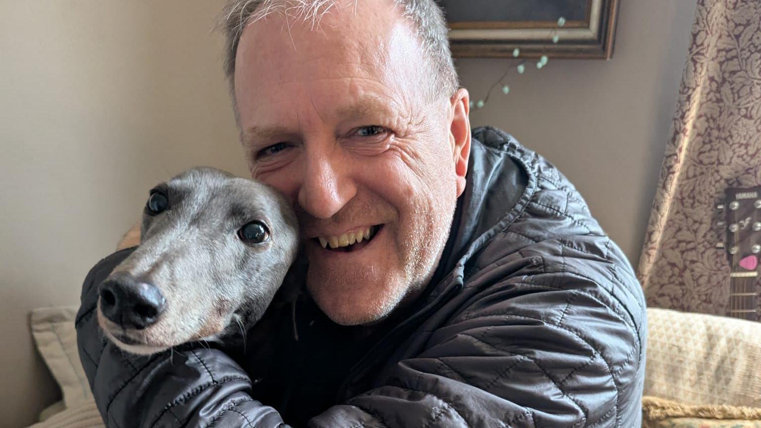 Essex and greyhound Gilly hug in his living room after the news that greyhound racing will be banned in Wales. Essex is wearing a black down jacket while Gilly is grey with a black nose.