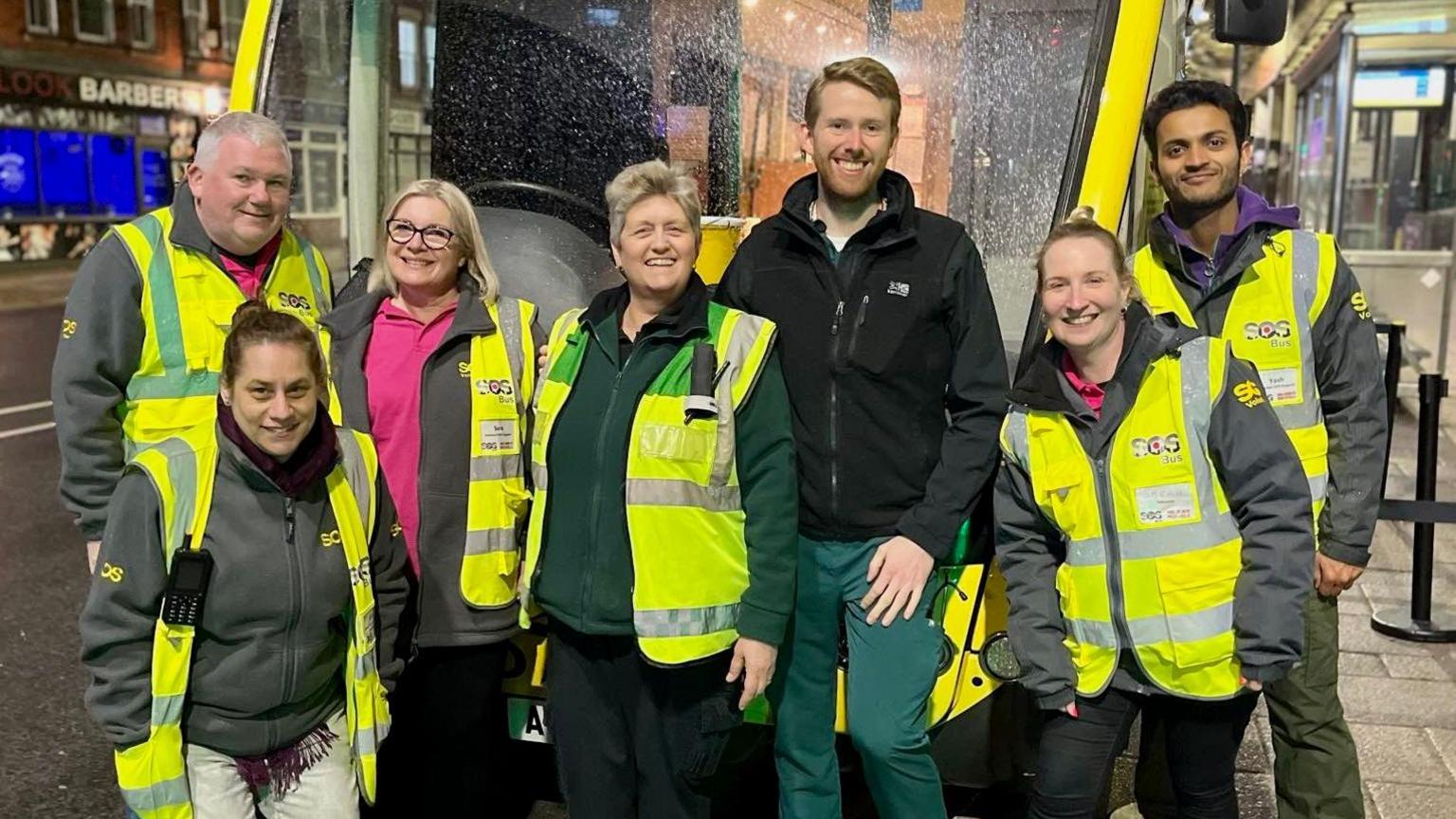 Volunteers and paid staff wearing yellow high-visibility vests are standing beside the SOS Bus