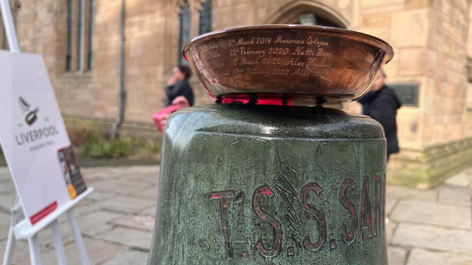 A close up of the copper frying pan with engraving of previous winners. The pan is on a bell in the grounds of the church with a board in the background saying Liverpool Pancake Race established 1982.