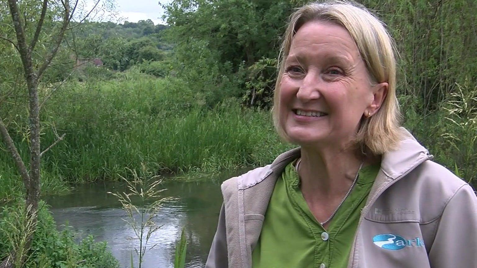 Charlotte Hitchmough stand infront of the riverbank, she is looking just off camera and smiling, has short blonde hair tucked behind her ears, wears a lime green buttoned blouse and beige unzipped jumper