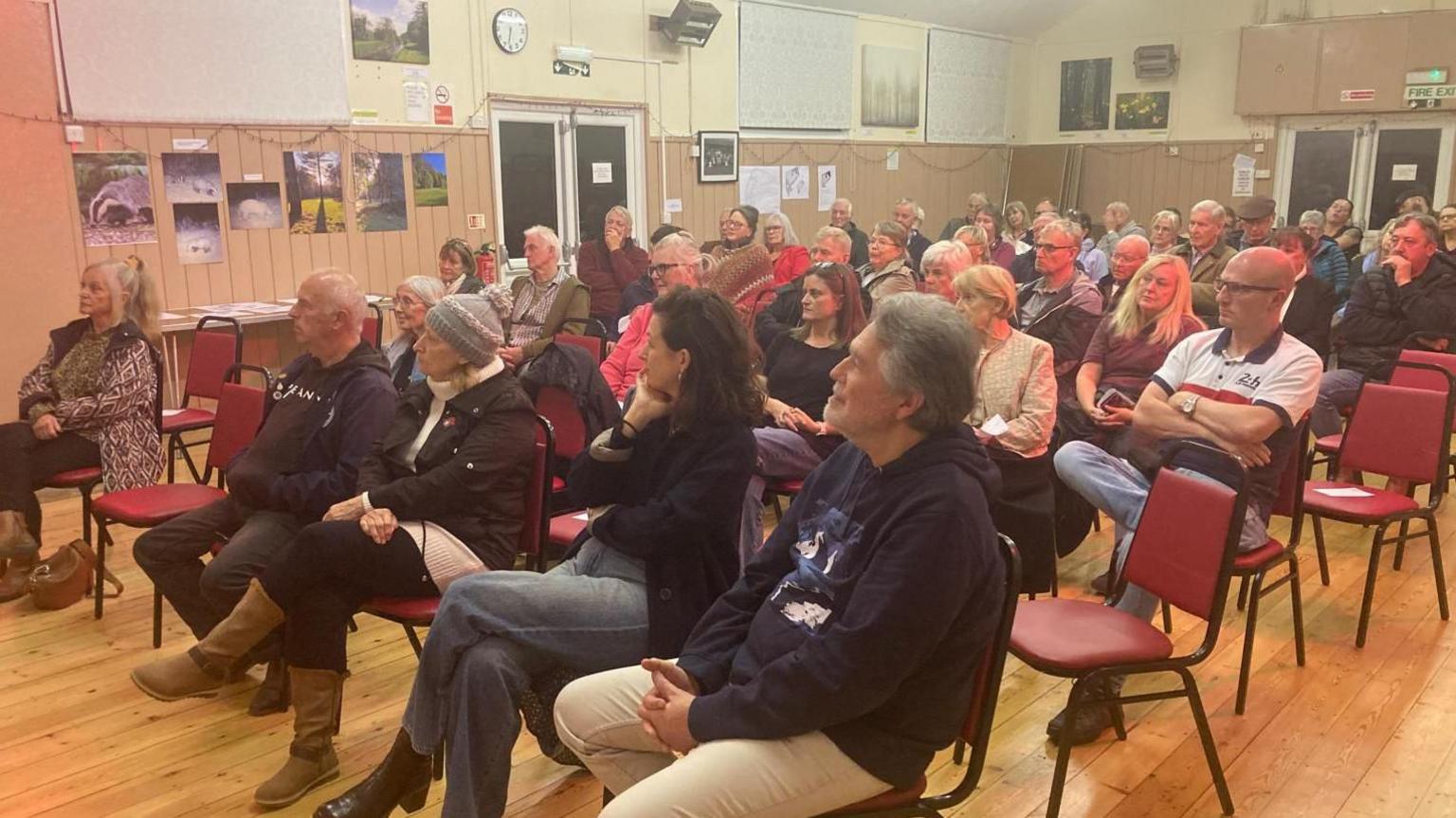 A group of people sat in rows of red chairs sitting in a village hall which has a wooden floor. There are about eight rows of chairs.